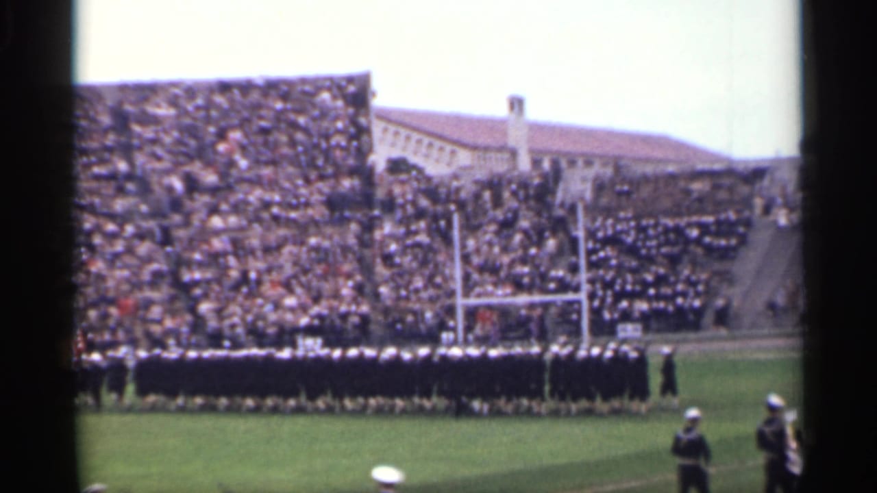Marching band in dark uniforms stands on a stadium field before crowded bleachers.