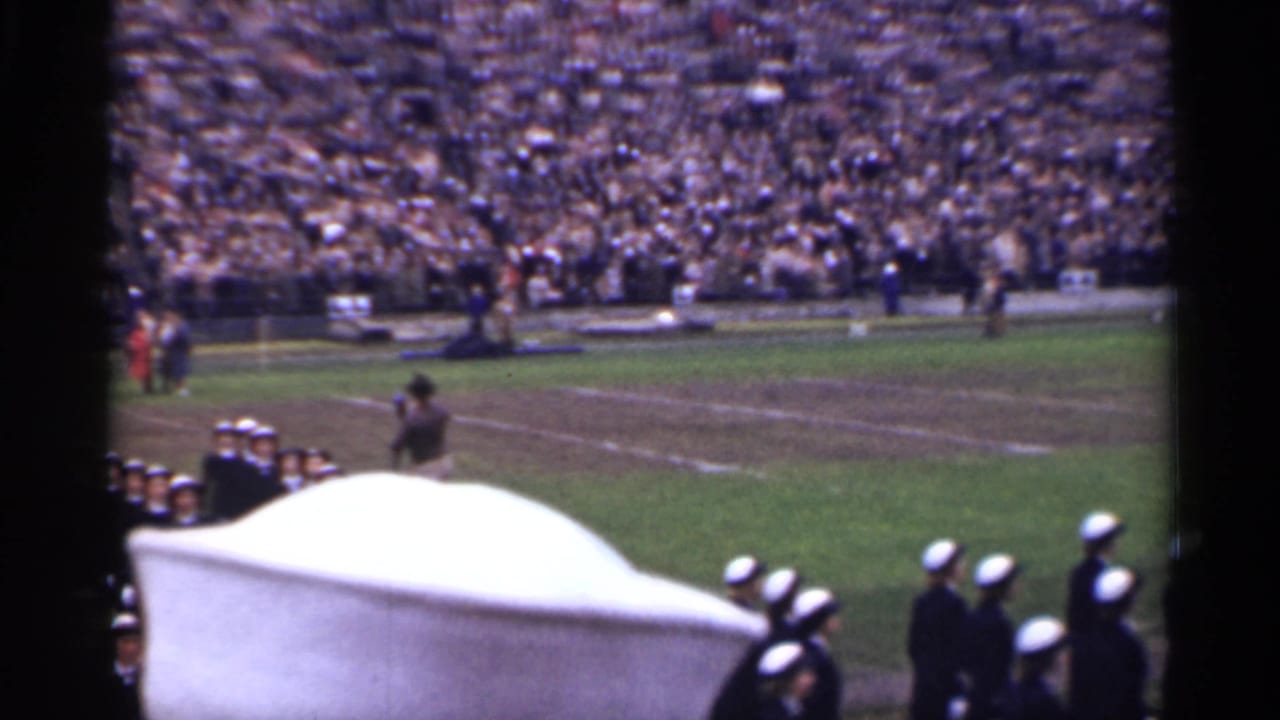 1944 home-movie frame of uniformed marchers on a stadium field, with spectators in the stands and a white object blocking part of the view.