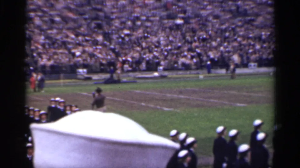 1944 home-movie frame of uniformed marchers on a stadium field, with spectators in the stands and a white object blocking part of the view.