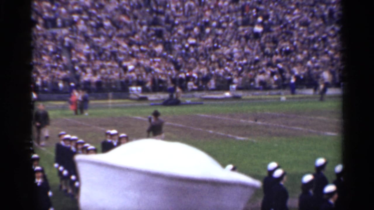 Soldiers and sailors gather near a football field as a large stadium crowd watches.