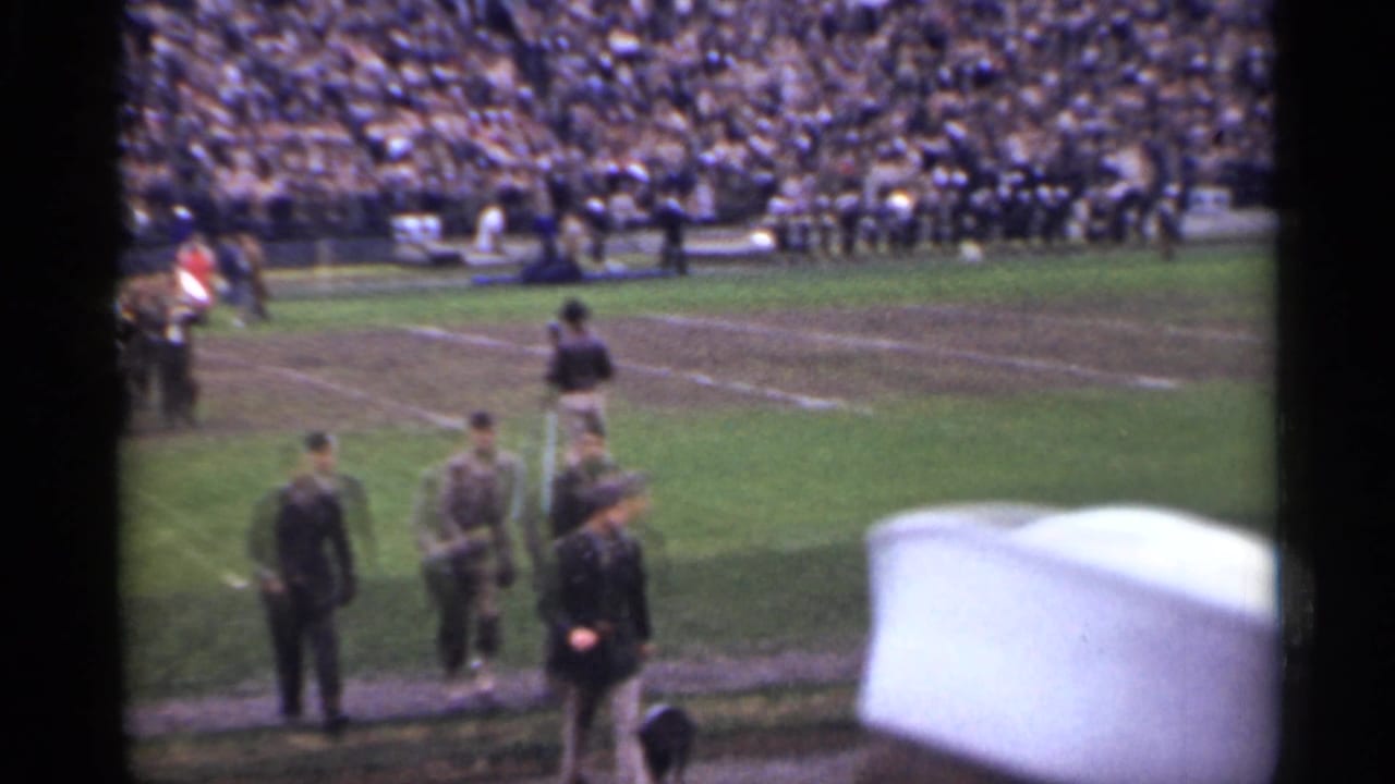 1944 San Francisco frame with soldiers and officials on a worn football field, viewed from behind a sailor's white cap in the stands.
