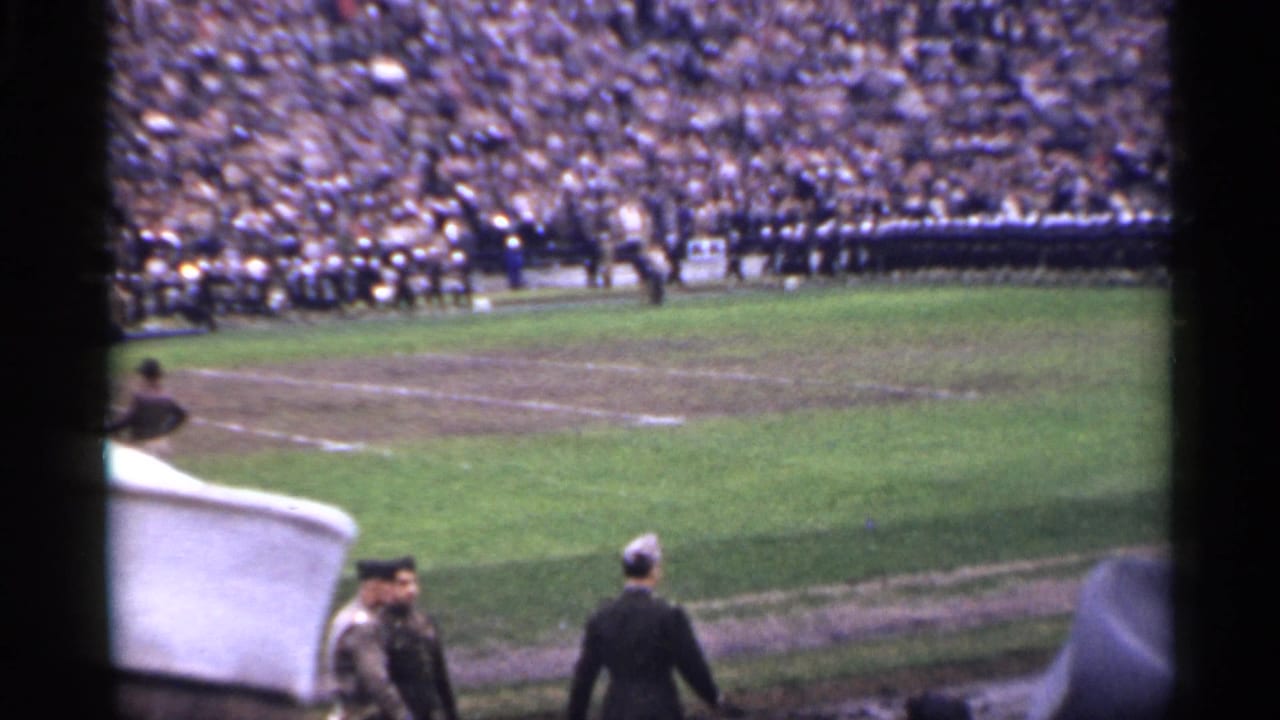 Packed stadium with uniformed formations on a field and people walking near the sideline.