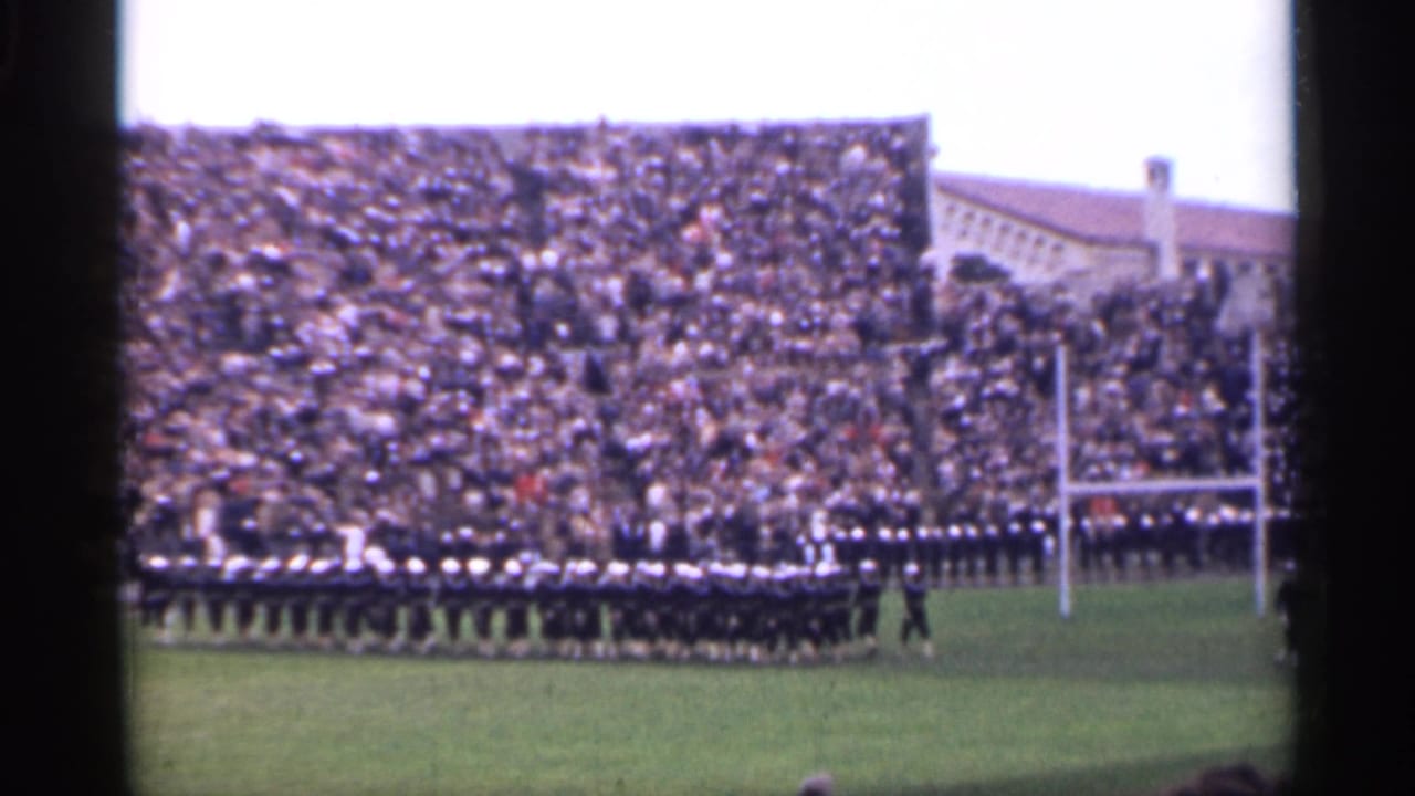 Wide home-movie frame of soldiers arranged across a stadium field, with goalposts and packed seating visible in San Francisco.
