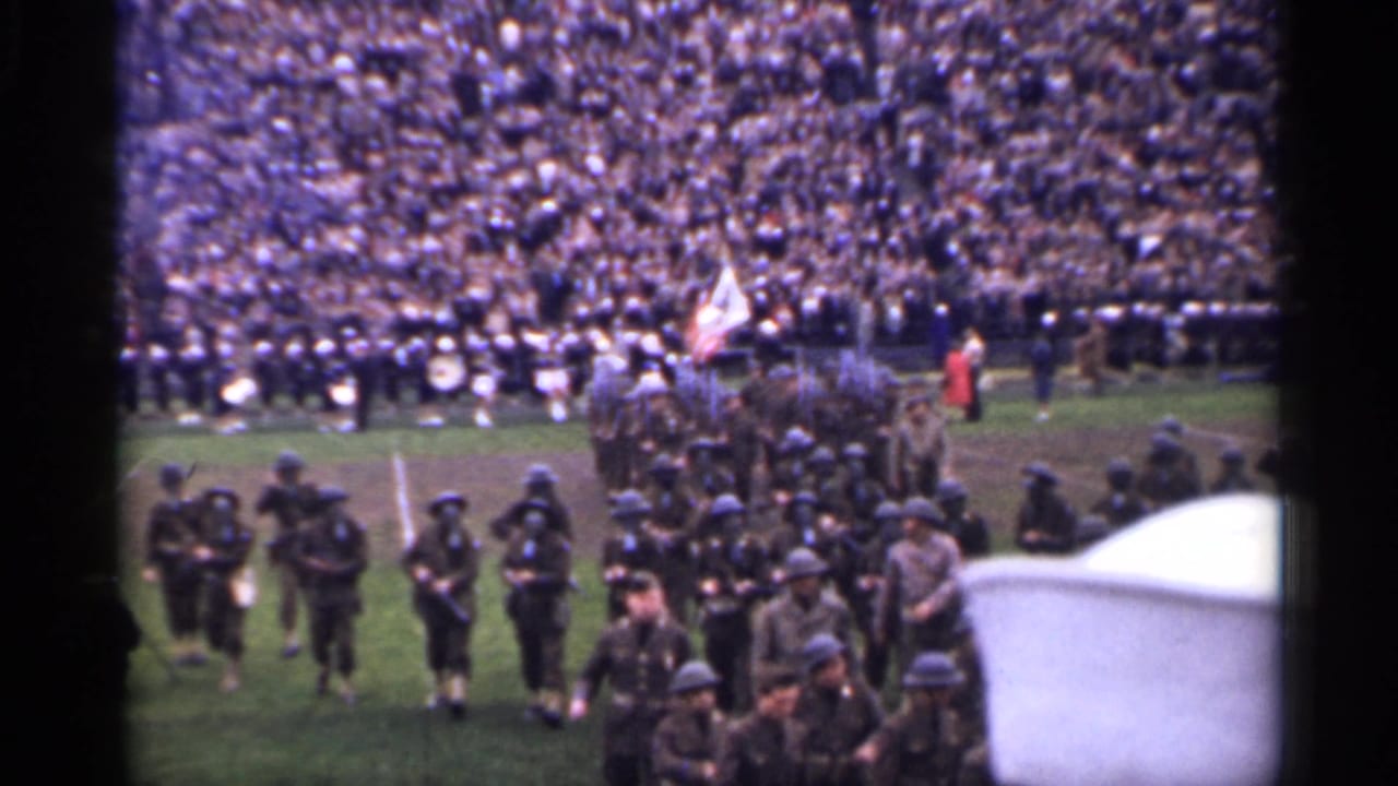 Soldiers and flag bearers march near the stadium track, with seated spectators and field activity visible behind them.