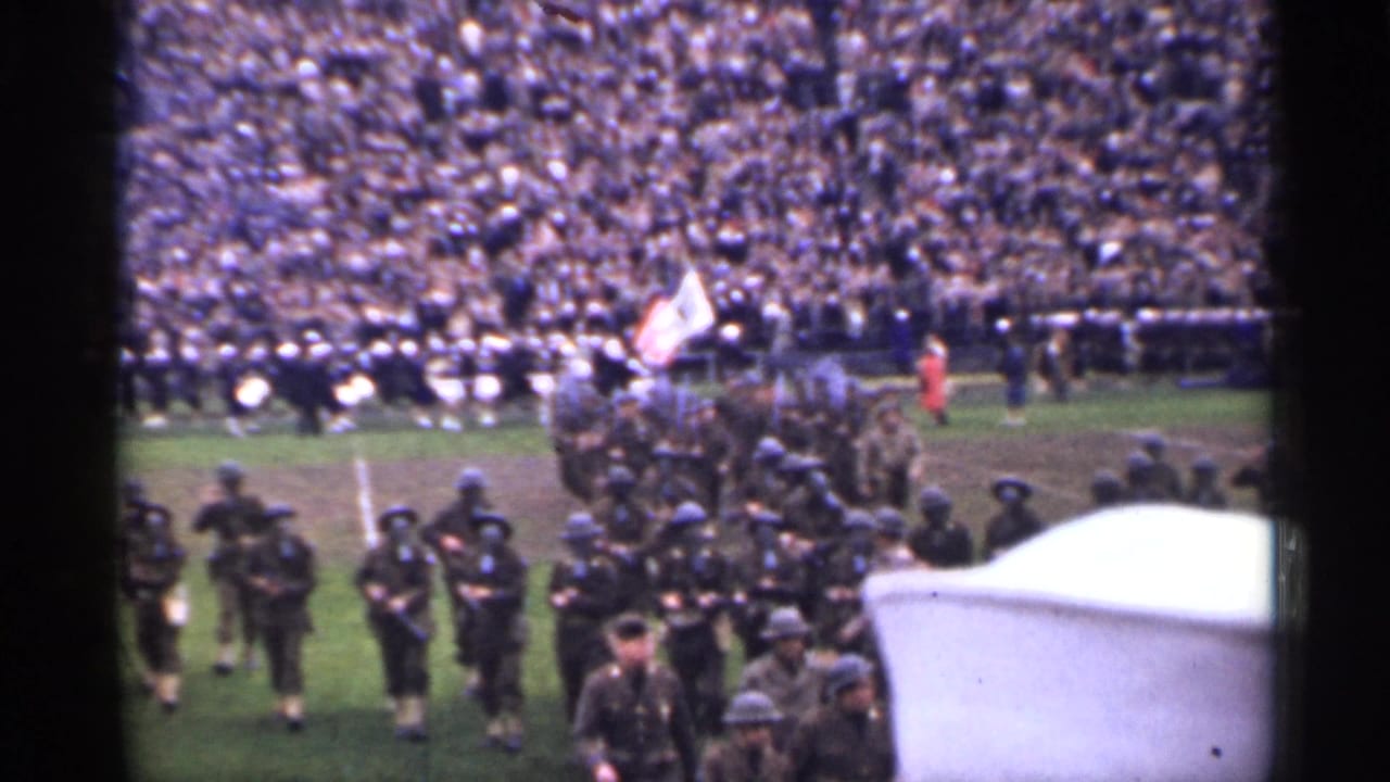 Soldiers march with rifles on a stadium field before a large crowd.