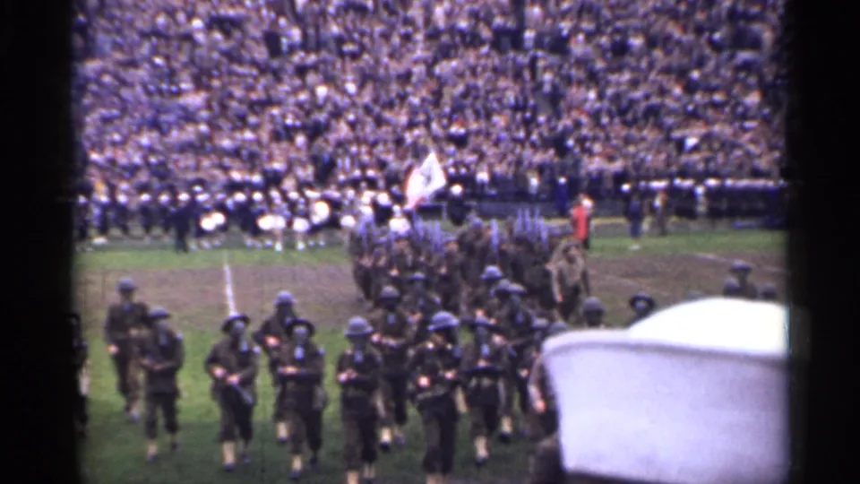 Uniformed marchers and a flag bearer cross a crowded San Francisco stadium field in 1944, with a band visible behind them.