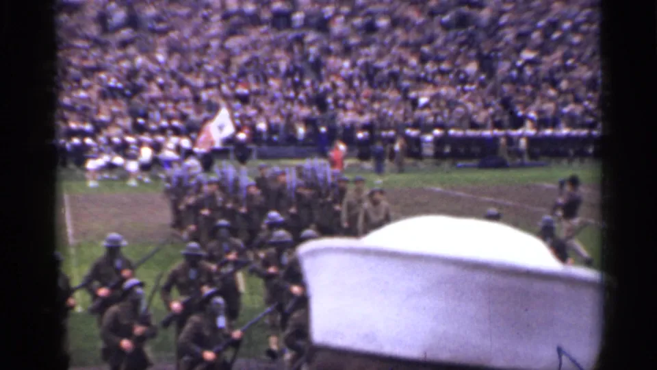 1944 San Francisco stadium scene with uniformed marchers moving past the camera, partly blocked by a foreground object and film gate edge.