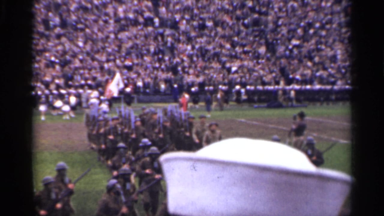Soldiers march across a stadium field as a crowd fills the stands and a white cap blocks part of the foreground.