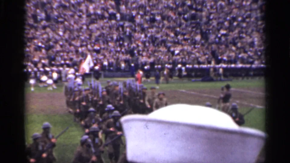 1944 San Francisco stadium view with soldiers marching in formation, spectators in packed stands, and a white sailor-style cap in the foreground.