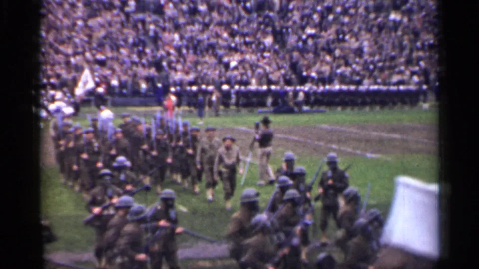 Soldiers in WWII-era uniforms march across a San Francisco stadium field before packed grandstands and visible flag bearers.
