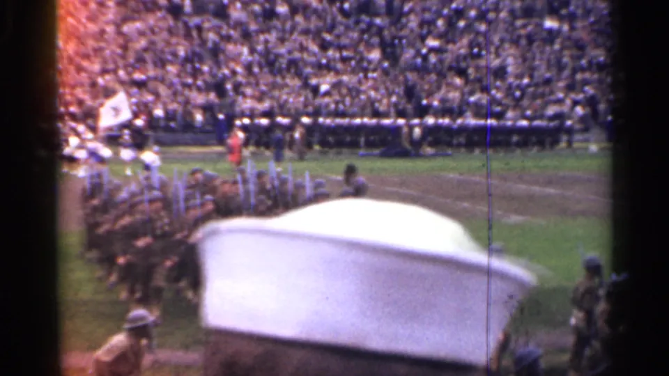 Stadium parade in San Francisco with marching military personnel, packed stands, and a large white sailor-style cap in the foreground.