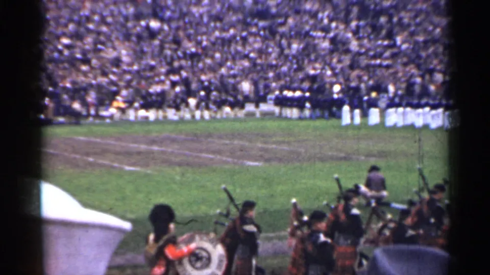 Home-movie frame of a San Francisco stadium performance, showing pipe band musicians and a bass drum before crowded stands.