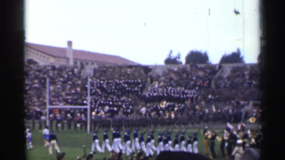 White-uniformed band members march across a 1944 San Francisco stadium field, with musicians and spectators visible behind them.