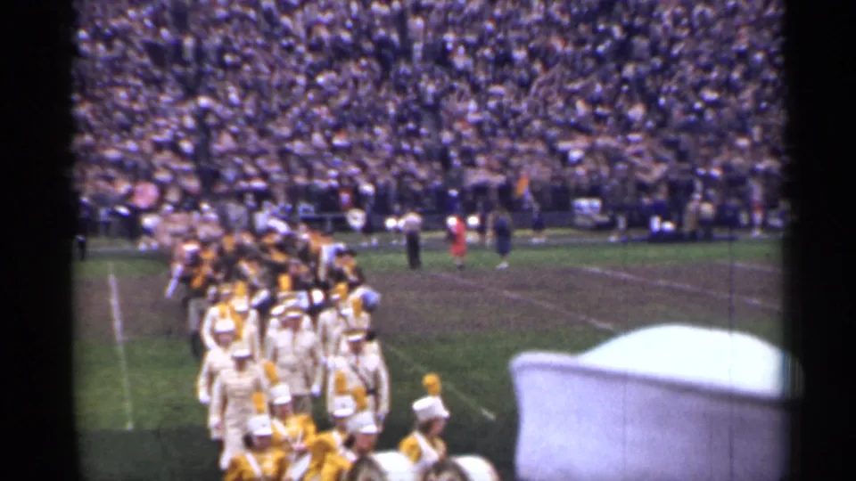1944 San Francisco football stadium frame showing a marching band crossing the field, partly obscured by a foreground sailor-style cap.