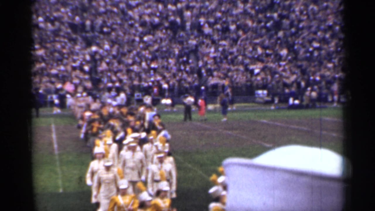 Marching band in light uniforms enters a football field before crowded stadium stands.