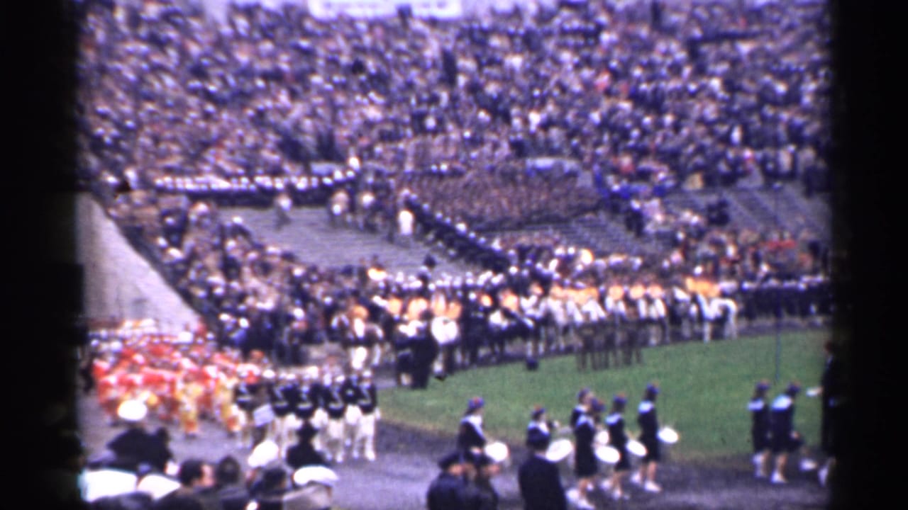 Uniformed band members march near the edge of a field with spectators behind.