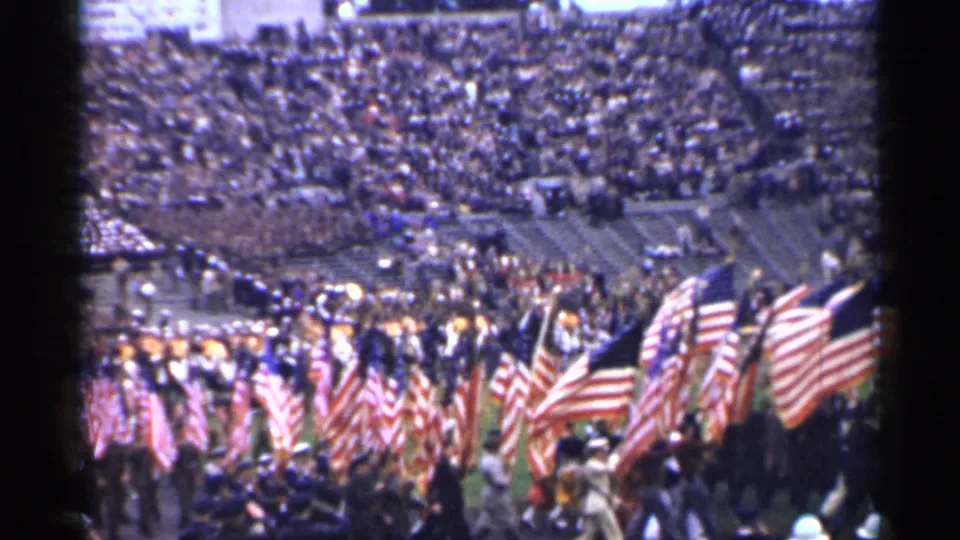 Home-movie frame of multiple American flags advancing across a stadium field in 1944 San Francisco, viewed from the stands.