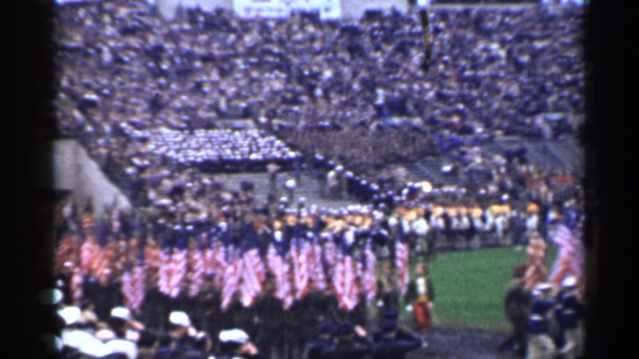 Flag bearers march across a stadium field before packed stands.