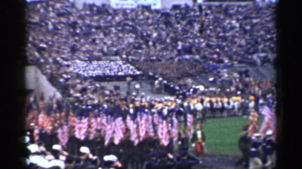 1944 San Francisco home-movie frame showing American flags and uniformed marchers entering a crowded stadium field.