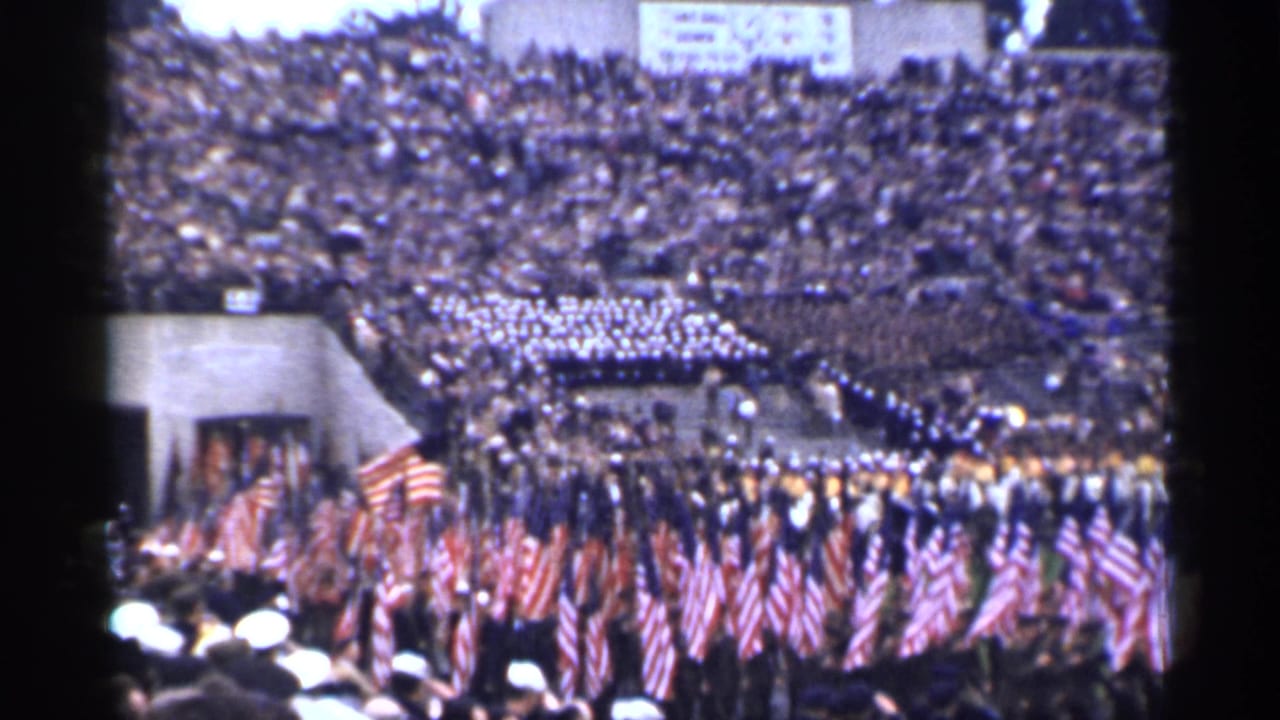 Crowded stadium with American flags and a band formation entering the field.