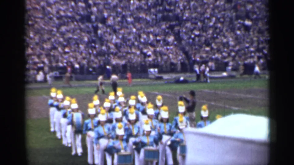 A marching band in light blue uniforms and yellow hats forms up near the sideline before a packed 1944 San Francisco stadium crowd.