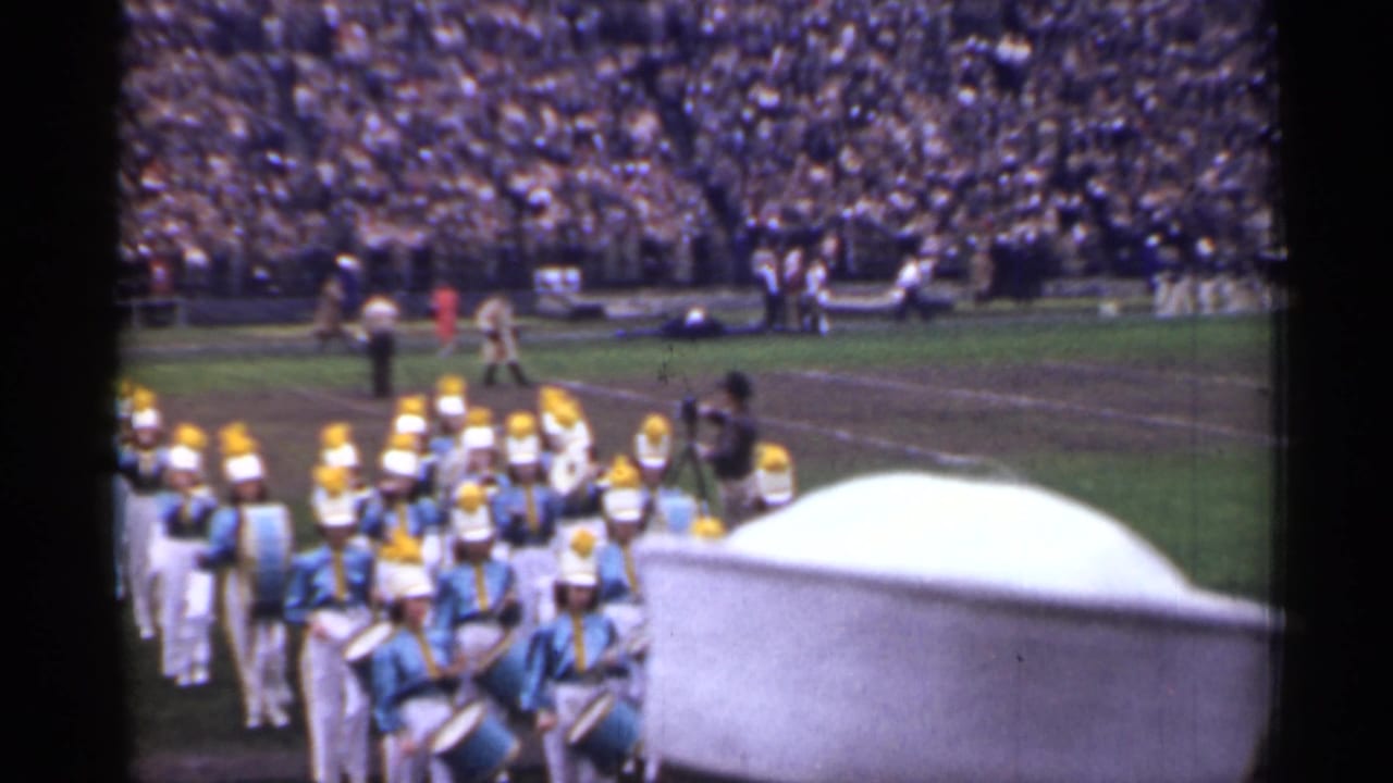 Marching band members in blue and white uniforms pass below crowded stadium stands.