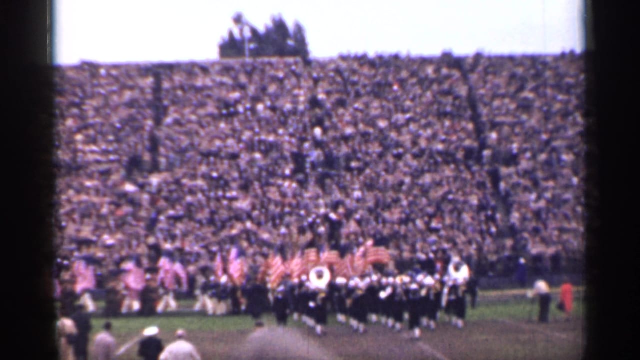 Marching band and American flags on a football field before packed stadium stands.