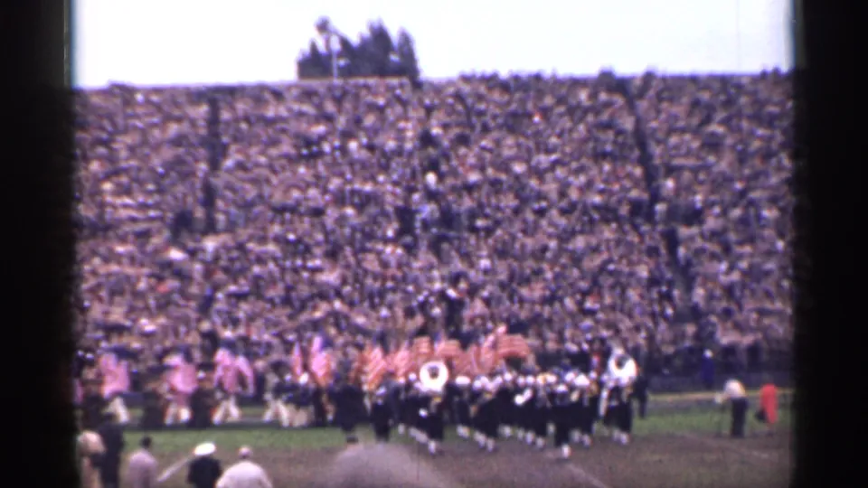 1944 San Francisco home-movie frame showing a marching band and American flags on a football field before crowded stands.