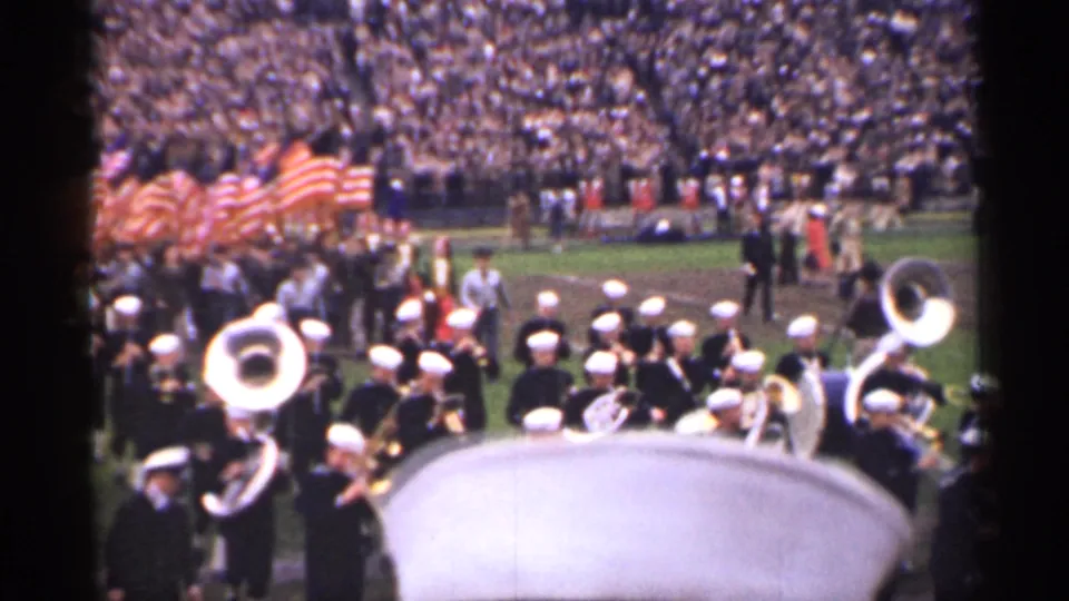 1944 San Francisco frame showing white sousaphones, uniformed band members, and clustered American flags near the sideline.