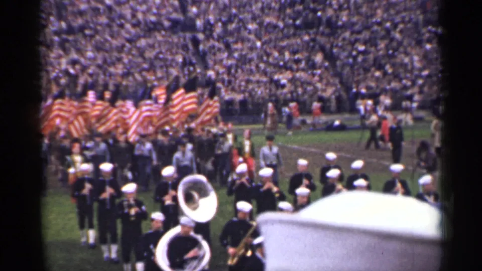 1944 San Francisco home-movie frame of a marching band, white sousaphones, and American flags beside a crowded football field.