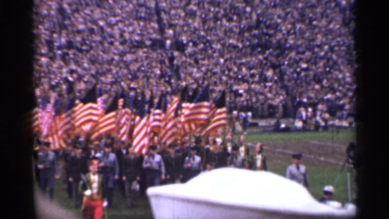 Uniformed marchers carry American flags past a packed stadium crowd with a white object in the foreground.