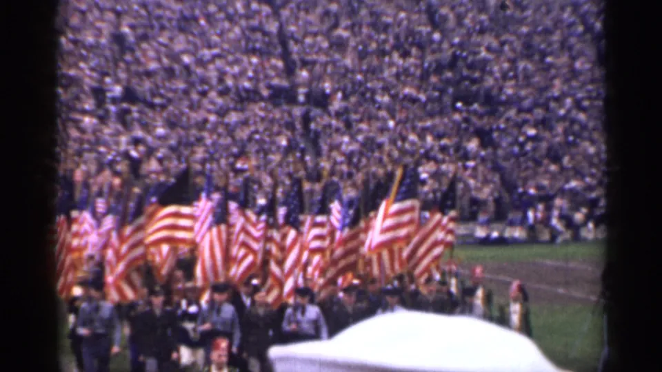 1944 San Francisco home-movie frame of a stadium parade with U.S. flags, uniformed marchers, and a large seated crowd.