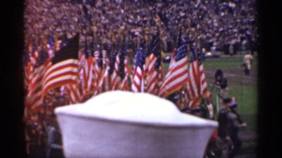 San Francisco 1944 parade frame with flags nearer the camera, uniformed marchers, a stadium crowd, and partial foreground blockage.