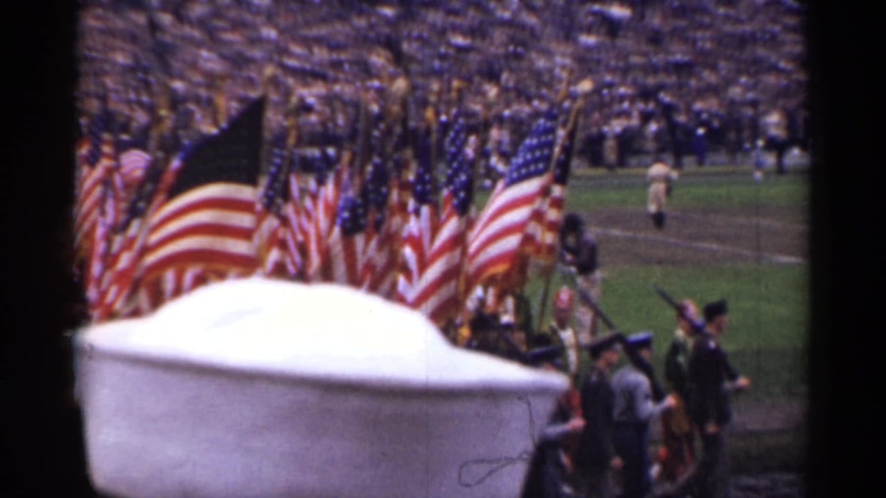 American flags and uniformed marchers pass before a large stadium crowd.