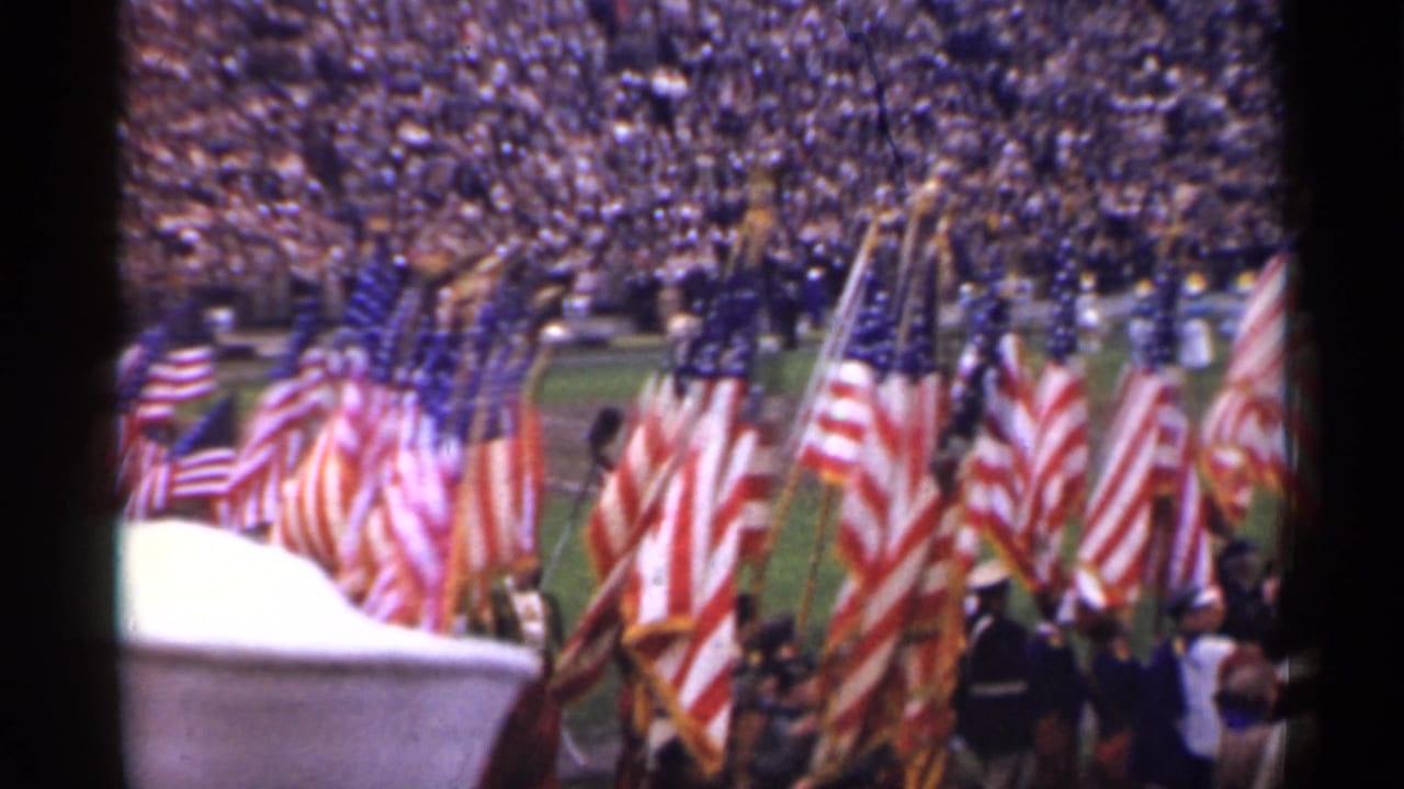 American flags carried on a stadium field before a large crowd.