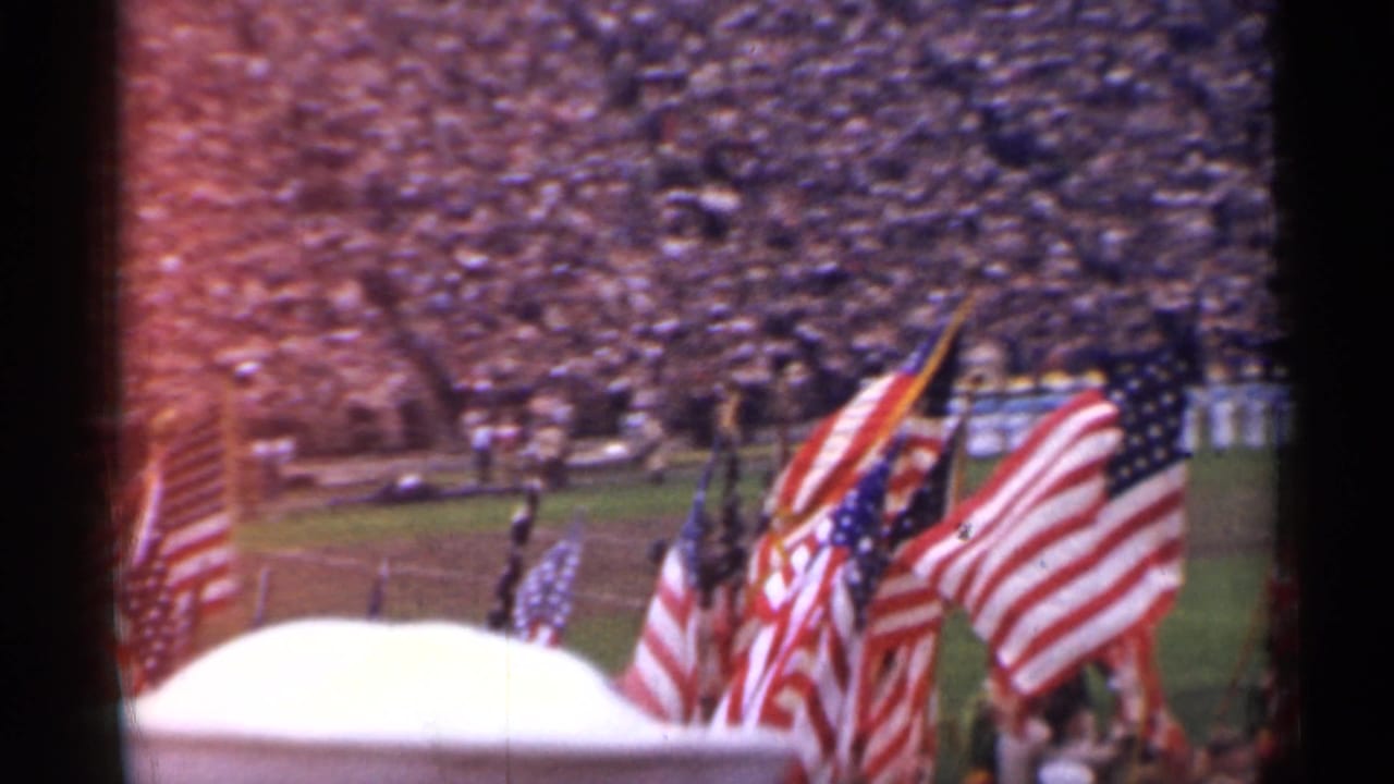American flags on a football field before a crowded stadium.