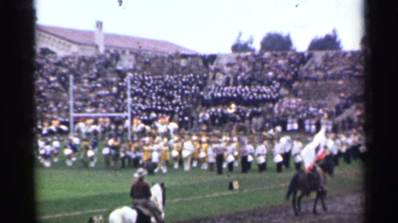 Marching band performs on a stadium field as horseback riders pass in front of crowded bleachers.