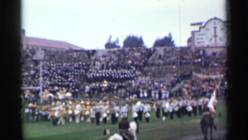 The stadium field shows a marching band in formation, horseback riders near the front, and dense spectator sections in the background.