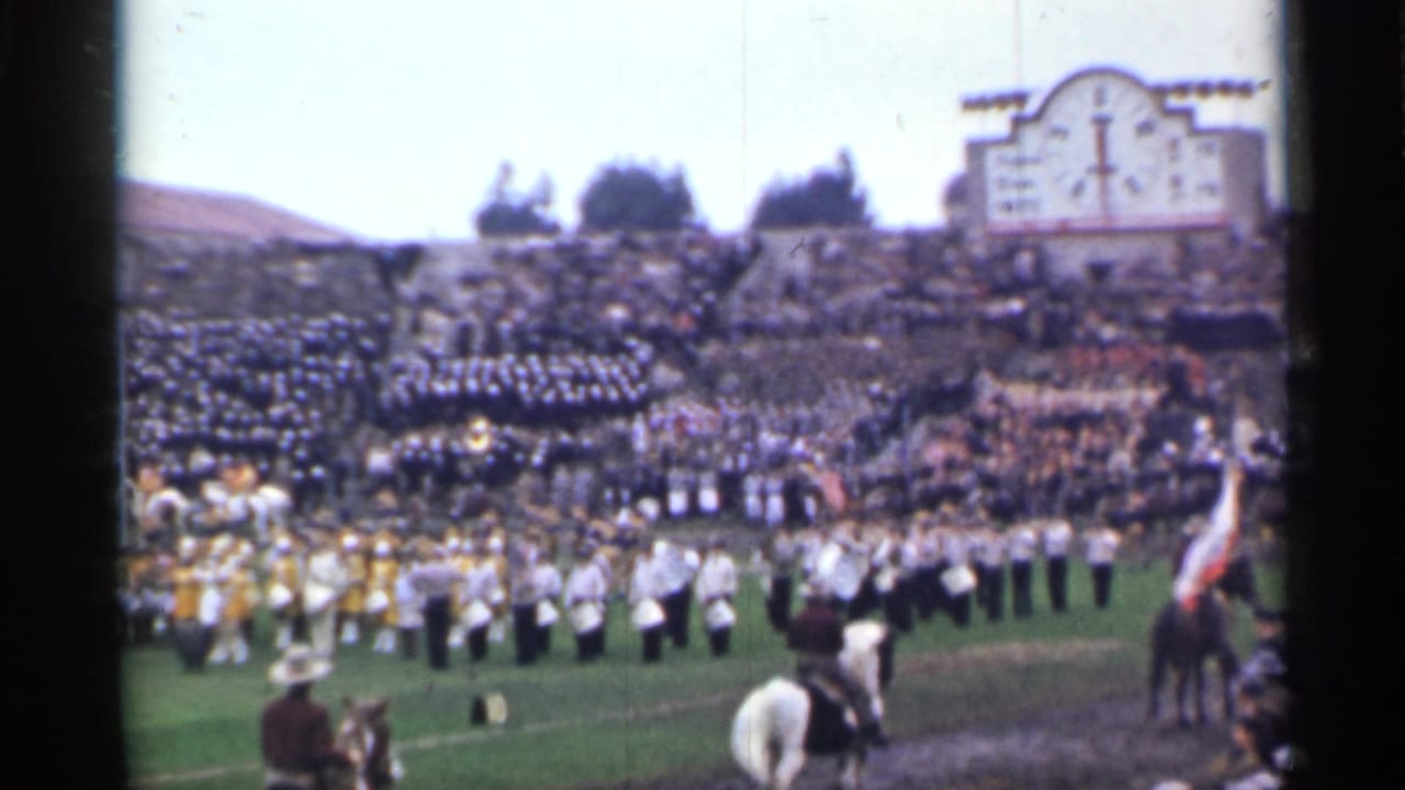 Crowded stadium with marching band on the field and mounted riders in the foreground.