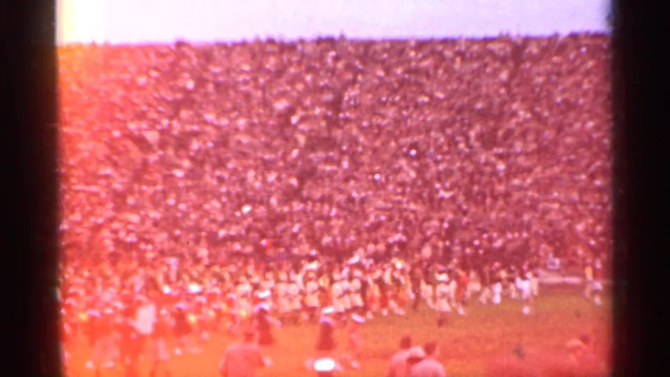 A color-shifted 8mm frame shows a large stadium crowd and performers lining the field during the 1944 San Francisco event.