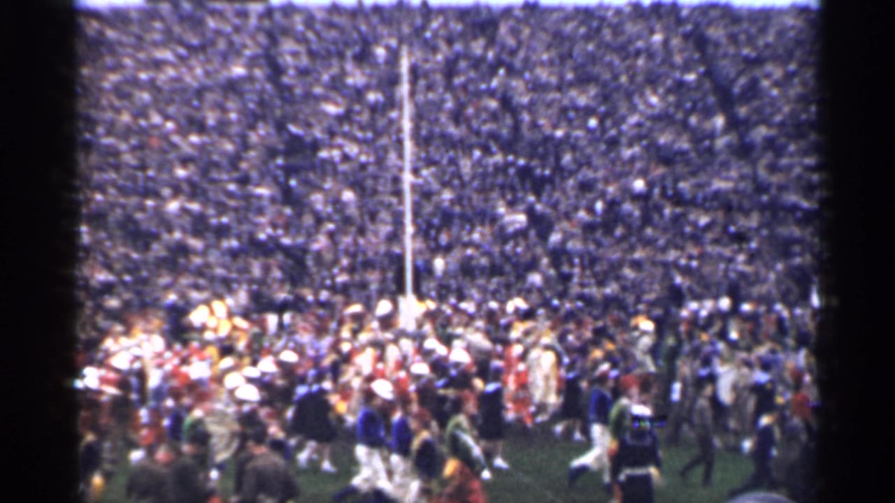 Packed stadium crowd behind a field with marching band members and a tall goalpost.