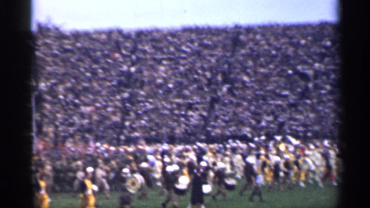 Marching band crosses a football field before packed stadium stands.