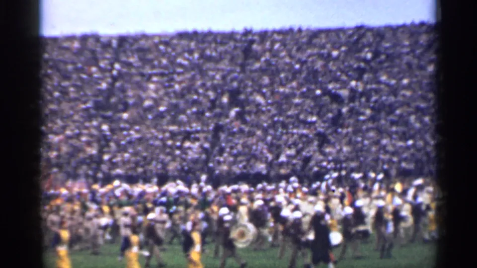 1940s 8mm footage frame of a marching band on a field below packed stadium stands, filmed from a distant spectator angle.