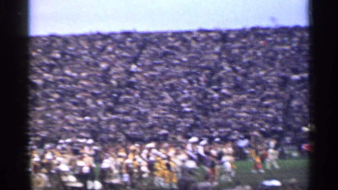 Crowded stadium stands with a marching band visible along the field edge.