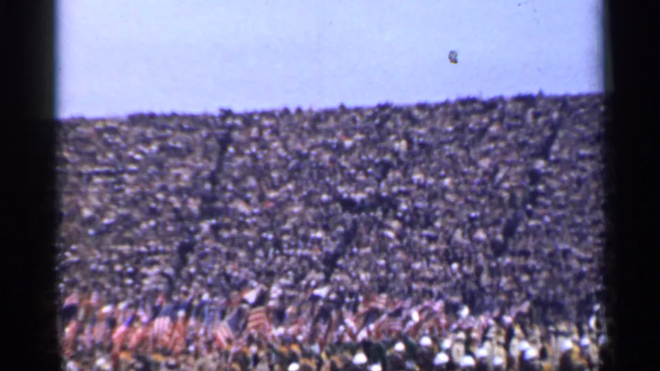 Wide 8mm frame of a 1944 San Francisco stadium gathering with flags and field-level performers.