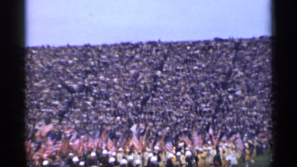 1944 San Francisco home-movie frame showing a dense stadium crowd behind a foreground group carrying American flags.