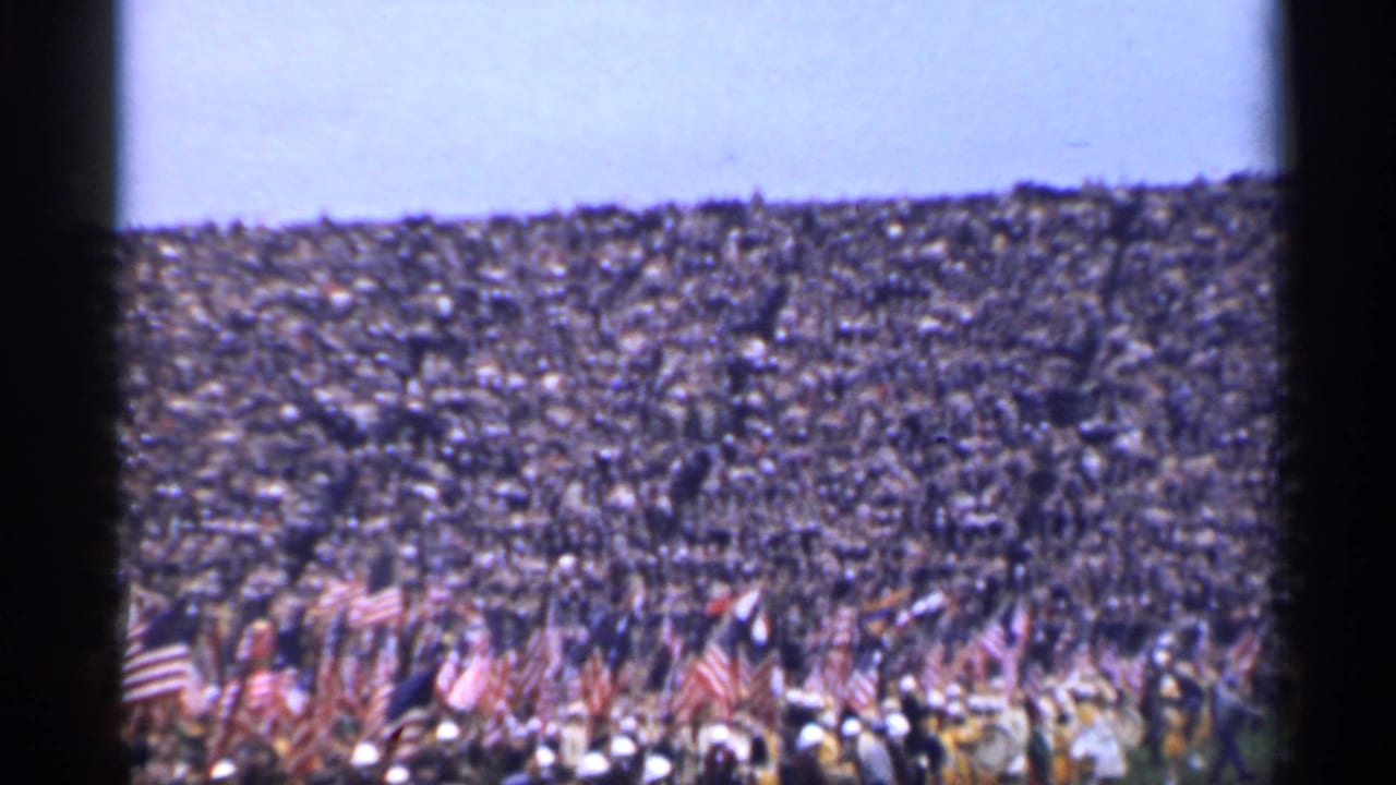 Distant crowd in stadium stands with many American flags at the front.