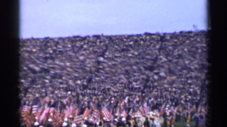 Wide 1944 home-movie frame of a packed public gathering in San Francisco, with flags in the foreground and stands beyond.