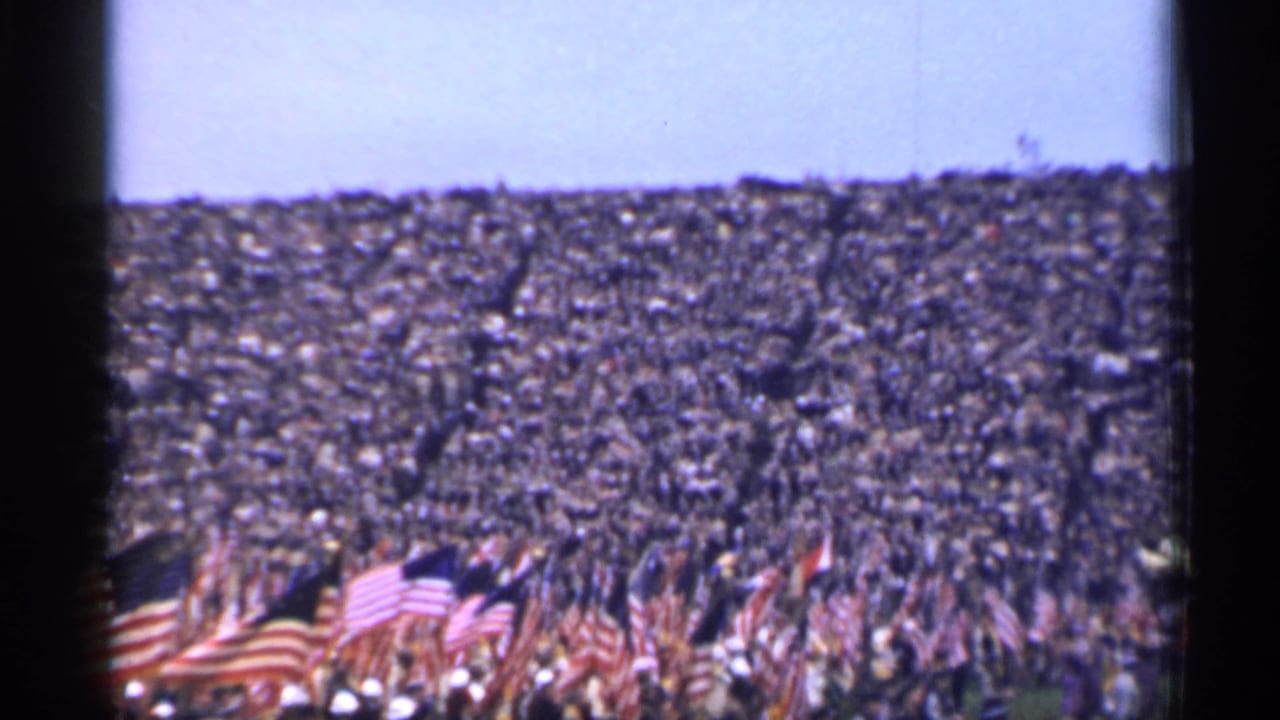 Packed stadium crowd with American flags waving in the foreground.