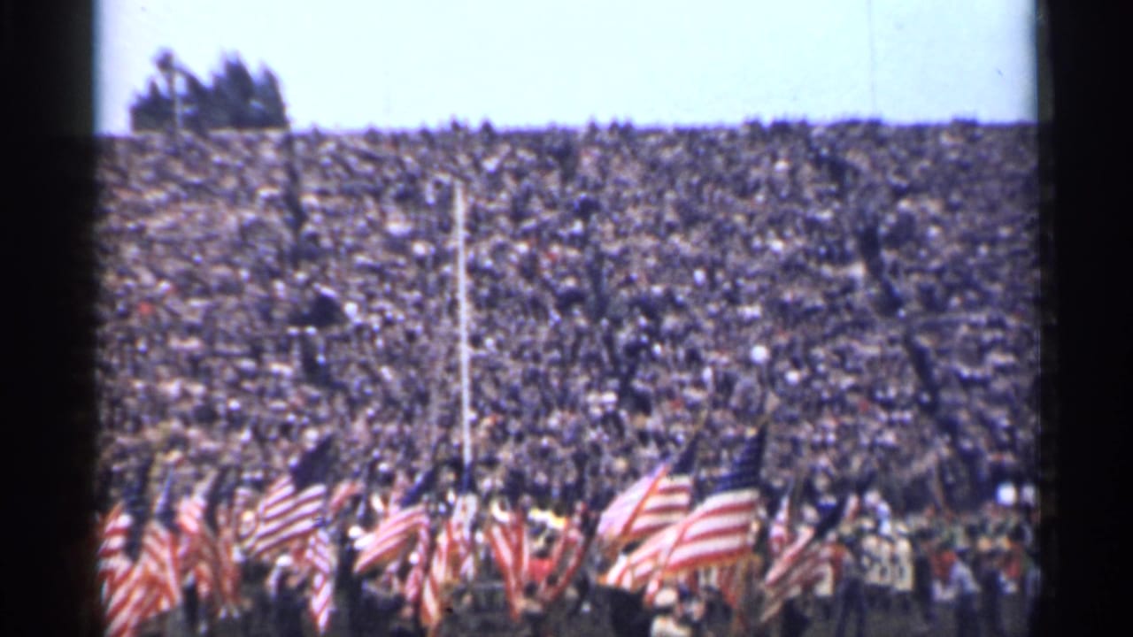 Packed stadium crowd with American flags and uniformed marchers in the foreground.